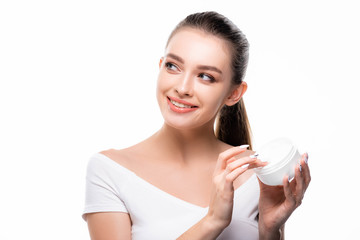 attractive, cheerful girl looking away while touching face cream in container isolated on white