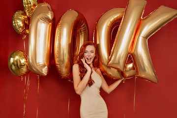 Girl with red hair wearing dress posing during photoshoot with confetti and balloons rejoices on red background, party, birthday, new year
