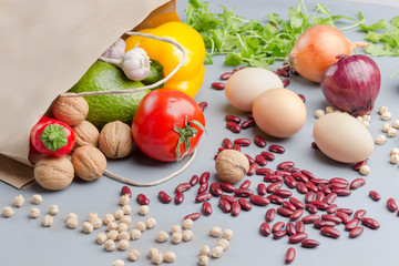 Obraz na płótnie Canvas A set of products for healthy eating. Paper bag with walnuts, chickpeas, beans, lentils, yellow and red peppers, tomatoes, avocado on a gray background.