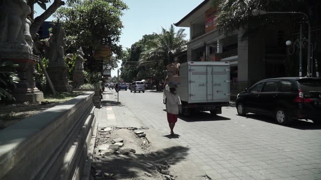 This is Balinese life in Ubud city, Bali. Daylife in the street with cars, motorcycles and people. Sunny day in summer, men working and caring stuff on his head. Filming handheld, steady shot
