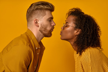 Mixed race couple stand in profile, keep lips folded, staring at each other, going to kiss, dressed casual mustard color wear, make grimace, pose over orange wall, have fun. Facial expressions concept