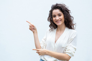 Joyful happy female customer pointing fingers away at copy space. Wavy haired young woman in casual shirt standing isolated over white background. Advertising concept
