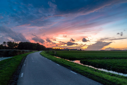 Road to sunset in Hazerswoude Rijndijk, the Netherlands. Country road during colourful sunset. Reflection in the small canals. Dutch meadows.