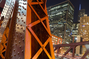 Night time detail shot of bridge overpass steel support columns with urban city skyline illuminated in background