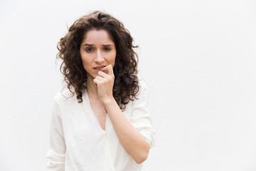 Pensive woman thinking and biting fingers and nails. Wavy haired young woman in casual shirt standing isolated over white background. Decision making concept