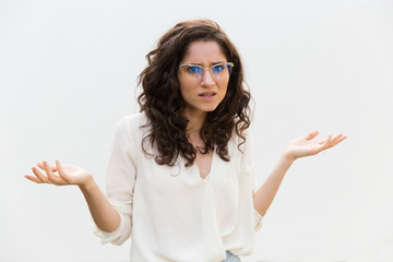 Puzzled annoyed woman in glasses shrugging and staring at camera. Wavy haired young woman in casual...