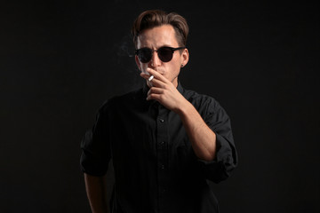 Fototapeta premium Stylish young man in a black t-shirt and sun glasses smoking a cigarette over black background.