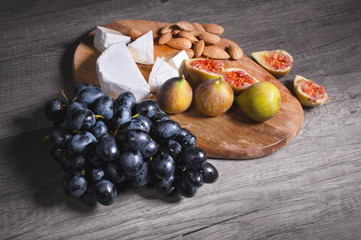Still life cheese plate on a wooden cutting board lies sliced cheese with mold almonds in the shell and sliced figs next to a bunch of black grapes. Delicious and healthy food concept