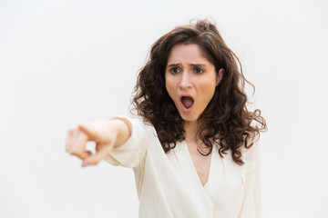 Shocked exaggerated woman with open mouth pointing finger away. Wavy haired young woman in casual shirt standing isolated over white background. Recognition or blaming concept