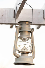 An imitation kerosene lamp under a retro silver color hangs on a wooden frame painted with white paint against a gray overcast sky.