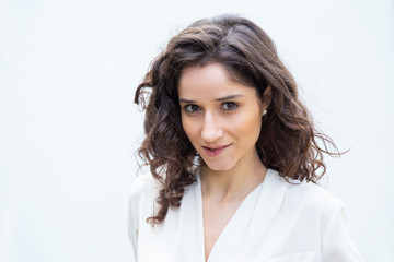 Cheerful sly beautiful student girl biting lower lip, looking at camera. Wavy haired young woman in casual shirt standing isolated over white background. Female portrait or flirt concept