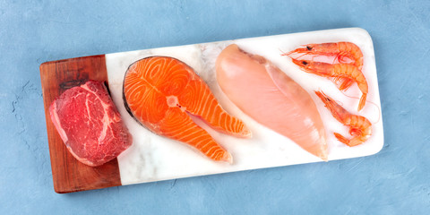 Fresh proteins panorama. Raw beef meat, salmon fish, chicken breast, and shrimps, shot from above