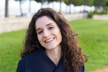 Closeup of smiling friendly beautiful student girl enjoying walking in park. Wavy haired young woman in casual overcoat posing outside. Leisure time in park concept