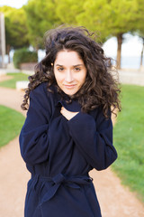 Friendly pensive beautiful student girl walking in park, feeling cold, wrapping herself in coat. Wavy haired young woman in casual overcoat posing outside. Autumn walk concept