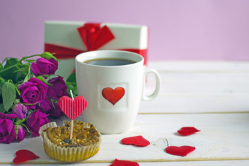 Valentine's Day breakfast. White tea mug with a heart pattern and  homemade cupcake with candles, hearts next to a bouquet of roses and a box with a gift on a wooden table. Copy space