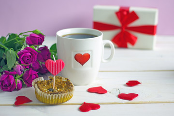 Valentine's Day breakfast. White tea mug with a heart pattern and  homemade cupcake with candles, hearts next to a bouquet of roses and a box with a gift on a wooden table. Copy space