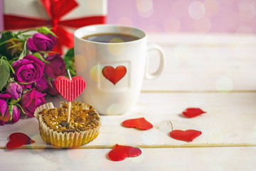 Valentine's Day breakfast. White tea mug with a heart pattern and  homemade cupcake with candles, hearts next to a bouquet of roses and a box with a gift on a wooden table. Copy space