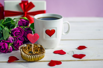 Valentine's Day breakfast. White tea mug with a heart pattern and  homemade cupcake with candles, hearts next to a bouquet of roses and a box with a gift on a wooden table. Copy space
