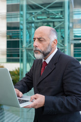 Focused mature businessman with laptop checking email near office building. Elderly man in formal suit and tie walking outside in city. Wireless technology concept