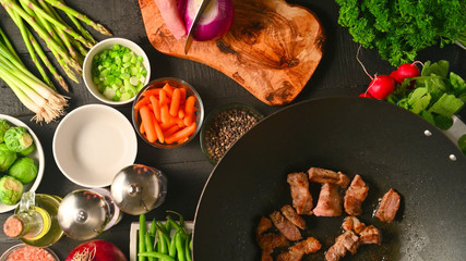 The meat is fried in a wok, against the background of which the cook slices onions. With various vegetables. Culinary background. Flat lay