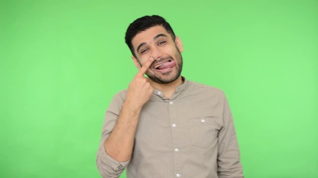 Funny positive brunette man with bristle in shirt picking his nose, pulling out boogers and making stupid grimaces with sticking out tongue, having fun. indoor studio shot green background, chroma key