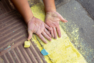 Child's hands face up stained with yellow and light blue chalk on a sidewalk, next to a yellow chalk and a light blue chalk, the sidewalk of the sidewalk painted yellow with chalk. Concept of play.