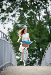 Young girl in a white summer dress is riding her blue citybike on the bridge, blurred trees on background, enjoying windy weather