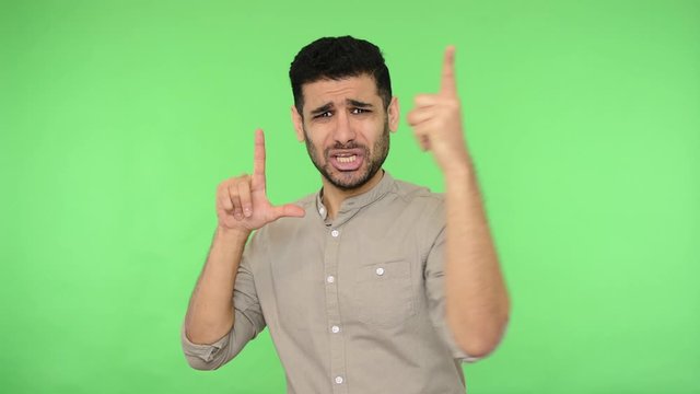 Angry brunette man with bristle in shirt showing loser gesture, L sign with fingers and pointing to camera, saying you are loser, blaming for defeat. indoor studio shot, green background, chroma key