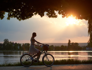 Obraz premium Happy woman riding a retro bike with a basket on the road near the lake on a sunset. Girl enjoying the magical view of the sun's rays of the falling sun at the end of the day