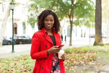 Woman with smartphone and paper cup smiling at camera. Beautiful young African American woman holding coffee to go and mobile phone while walking outdoors. Technology concept