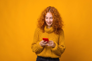 Photo of cheerful young redhead woman using her smartphone over yellow background