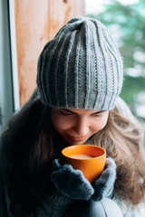 Beautiful  woman holding and drinking a cup of coffee or cocoa in gloves sitting home by the window. Blurred winter snow tree background. Morning, coziness and people concept