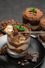 Classic tiramisu dessert in a glass cup and pieces of chocolate on stone cutting board on dark concrete background