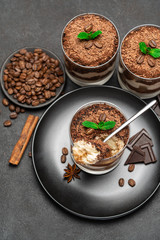Classic tiramisu dessert in a glass cup and pieces of chocolate on stone cutting board on dark concrete background