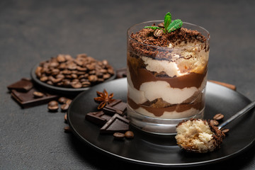 Classic tiramisu dessert in a glass cup and pieces of chocolate on dark concrete background