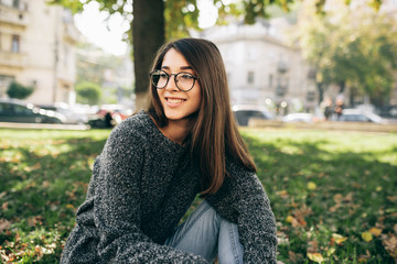 Outdoor image of beautiful smiling young woman sitting on green grass in the city park, wearing knitted sweater and transparent eyeglasses. Beautiful young woman resting outside