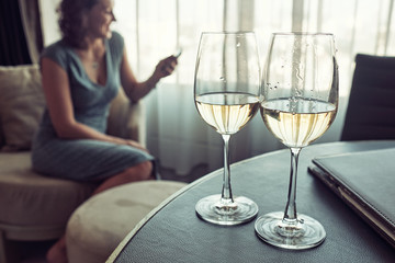 Two glasses of white vine on the table, on the background cheerful woman siting on the chair and using smartphone.
