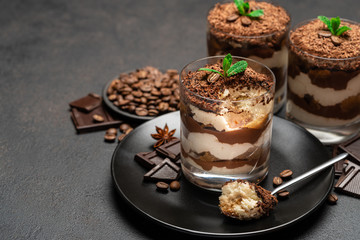 Classic tiramisu dessert in a glass cup and pieces of chocolate on dark concrete background