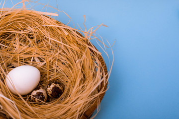 White chicken and quail eggs are in a wicker basket with straw on  blue background. Easter card greeting. Flat lay. Copy space.