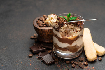 Classic tiramisu dessert in a glass cup and pieces of chocolate on dark concrete background