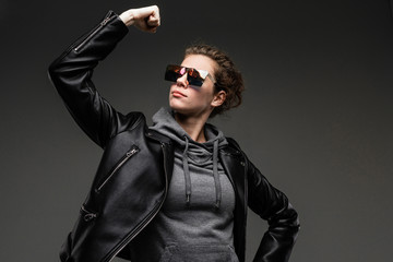 A young girl with rough facial features in a black jacket shows her muscles isolated on black background