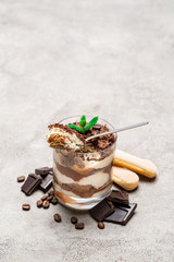 Classic tiramisu dessert in a glass cup, savoiardi cookies and pieces of chocolate on concrete background