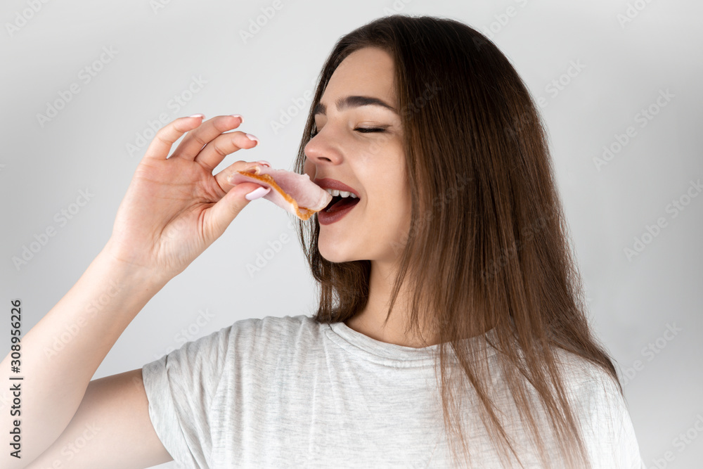 Wall mural young beautiful woman biting slice of ham in tempting manner looking hungry isolated white background