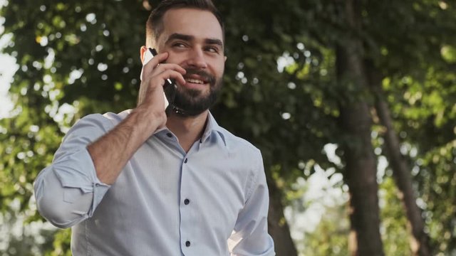 Happy bearded businessman answering the call and talking by smartphone while sitting in park outdoors