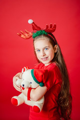 Portrait of a funny cheerful girl with a bandage of horns on her head hugging a teddy bear in Christmas pajamas isolated on a bright red background.