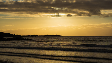 A lighthouse in the distance. The sun shining down from above, and in the foreground ocean waves rolling in on the beach. Gimsøya, Lofoten Islands, Norway.