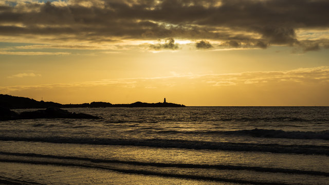 A lighthouse in the distance. The sun shining down from above, and in the foreground ocean waves rolling in on the beach. Gims&oslash;ya, Lofoten Islands, Norway.