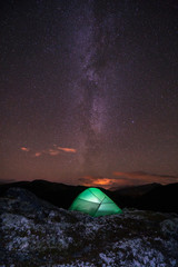 Milky way and starry sky over night scene outdoors in the forest and the mountains with green tent infront. Landscape, astronomy and camping concept. Vertical shot.