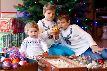 Two little kid boys and adorable baby girl decorating Christmas tree with old vintage toys and balls. Family preaparation celebration of family feast. Children, brothers and sister in festive clothes.