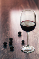 A glass of red wine on a wooden table with a glint from the window. Grapes are scattered on the table. Vertical orientation